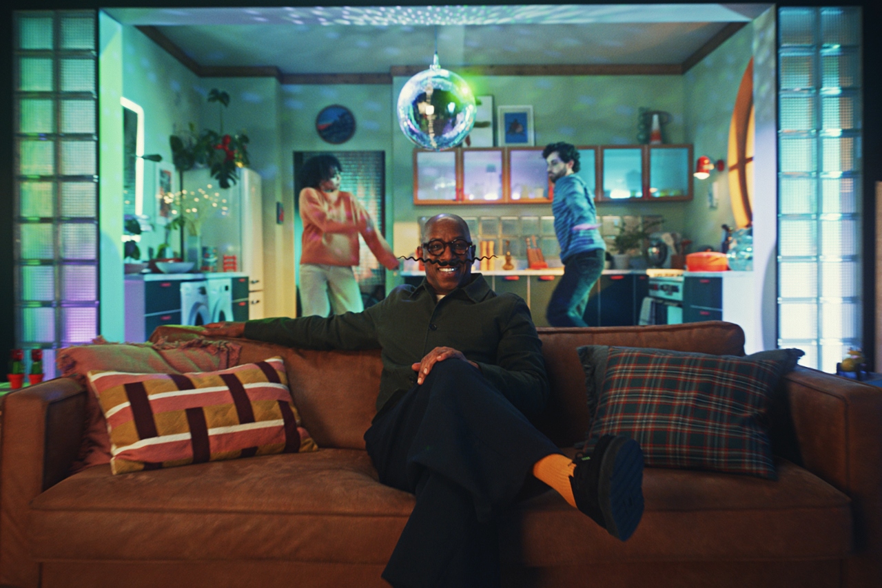 Ian Wright sits on a sofa smiling at the camera as a couple dance in their kitchen behind him. Wright is sporting a curly handlebar moustache and the couple's kitchen is lit by a disco ball.