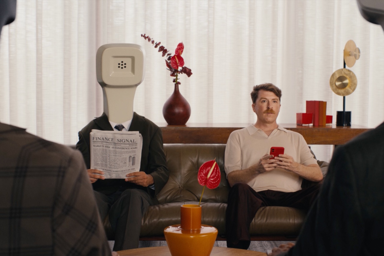 Person with telephone for a head sat on sofa next to man holding a mobile phone