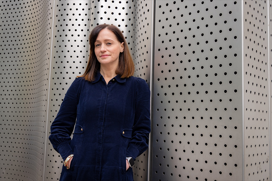 Victoria Appleby standing against a perforated metal wall, hands in pockets, wearing navy.